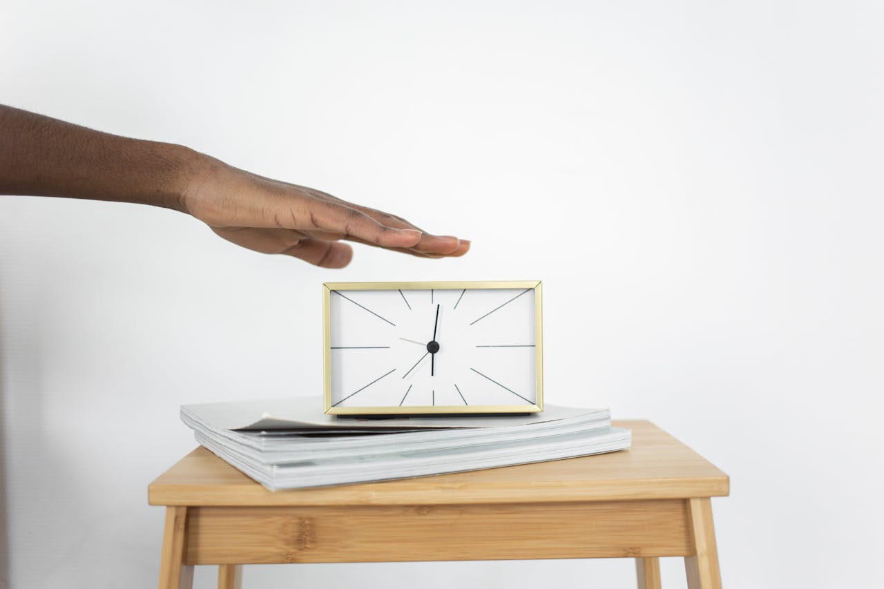 A hand reaching towards a minimalist alarm clock on a wooden table with books.